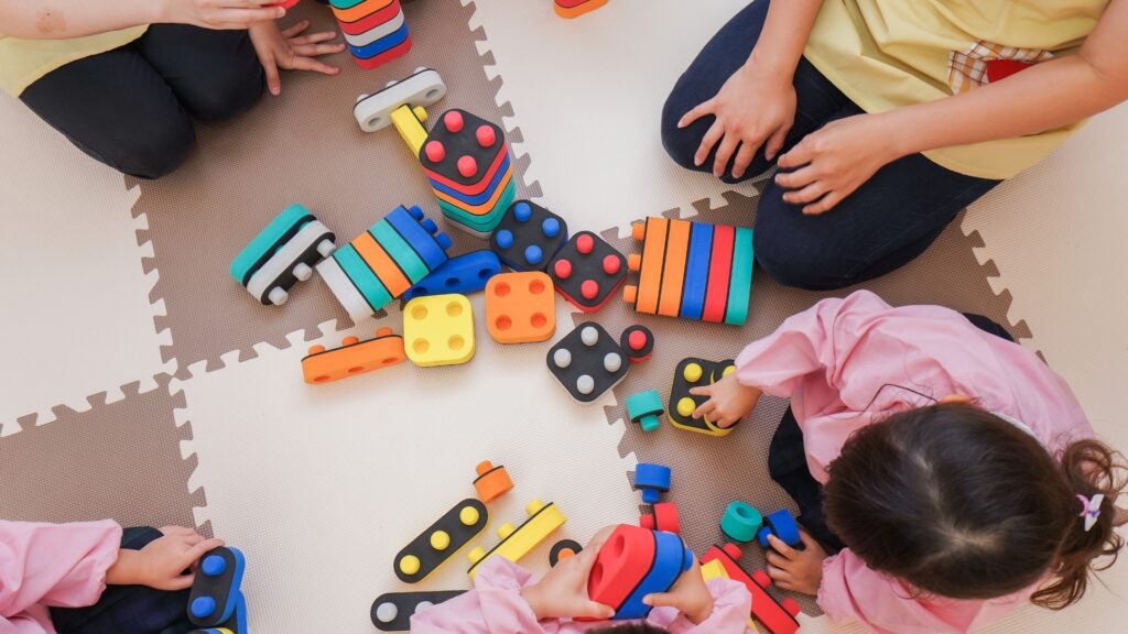 Children sitting on foam puzzle mats, playing with colorful stackable blocks and toy vehicles scattered on the floor