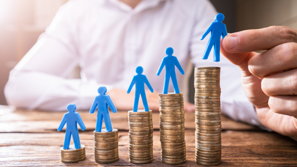 Blue figurines stand on ascending coin stacks, symbolizing financial growth and teamwork, with a hand placing the top figure on the tallest stack.