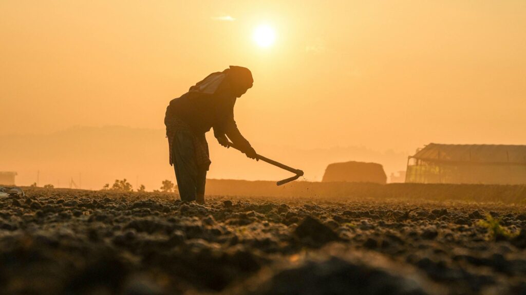 Silhouette of a worker digging with a hoe in a dusty field at sunset, with an industrial building in the distance.
